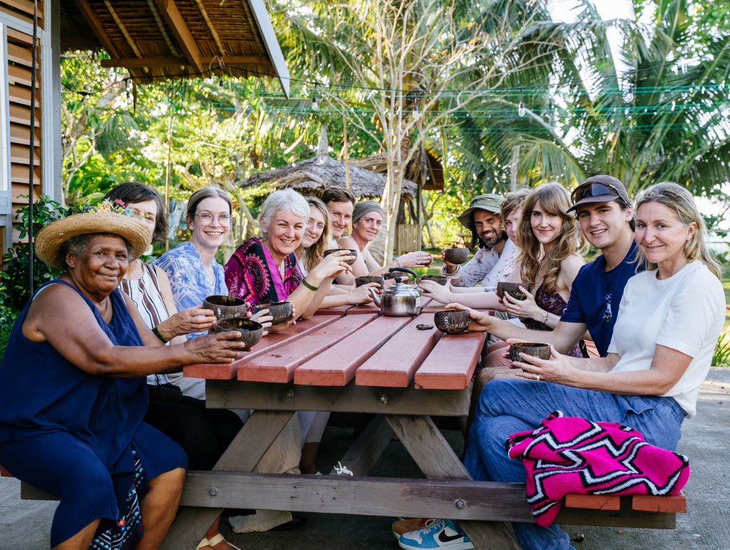 Group of people sitting around a wooden picnic table outdoors with greenery in the background at a cacao farm