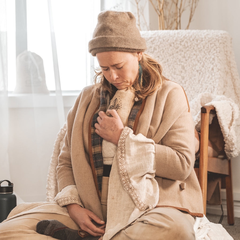 Woman sitting on a chair wearing a beige coat, plaid scarf, and brown hat in a cozy indoor setting.