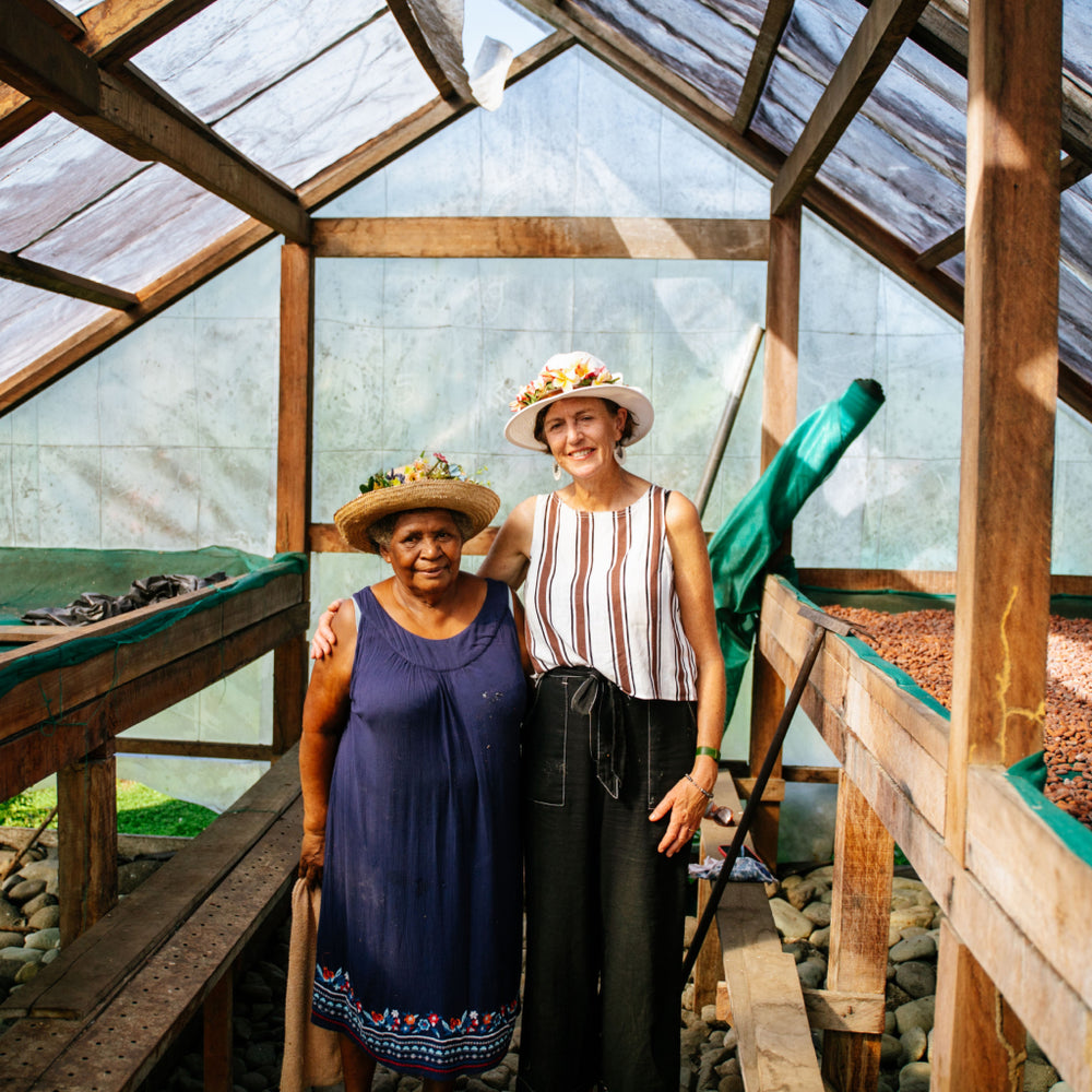 Two women standing inside a greenhouse with wooden benches and cacao.