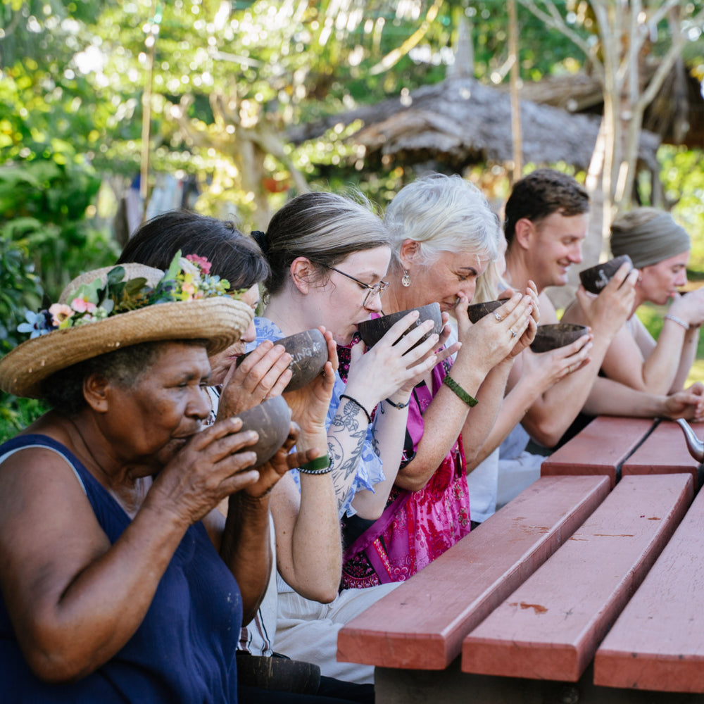 Group of people drinking from cups outdoors with a wooden table and greenery in the background.