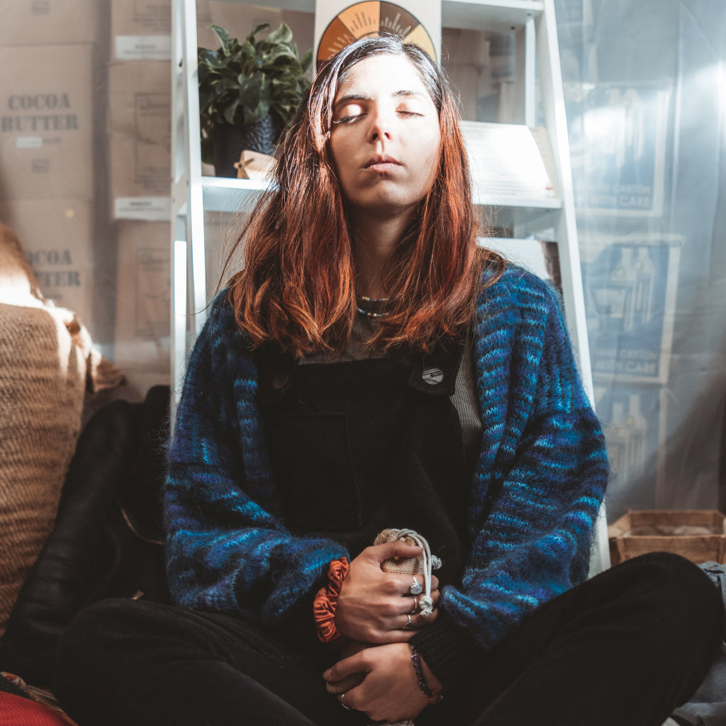 Person wearing a blue sweater sitting in a room with shelves and plants.