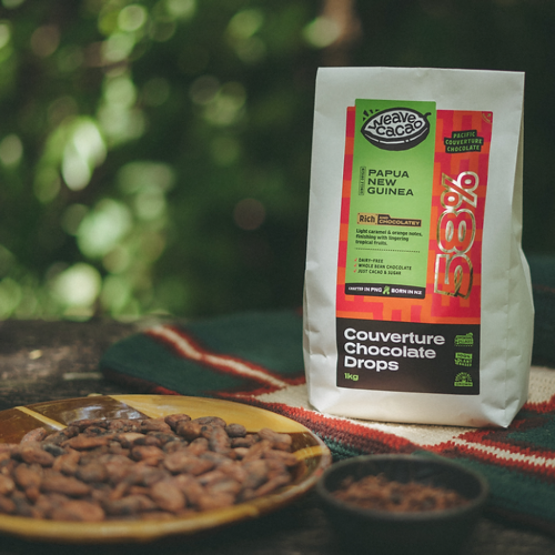 A package of Weave 58% couverture chocolate drops with a plate of cocoa beans in the foreground, set on a wooden surface with a greenery background.