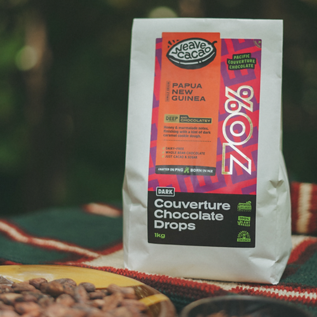 A package of Weave 70% Couverture Chocolate Drops with a colorful label, resting on a fabric next to raw cacao beans.