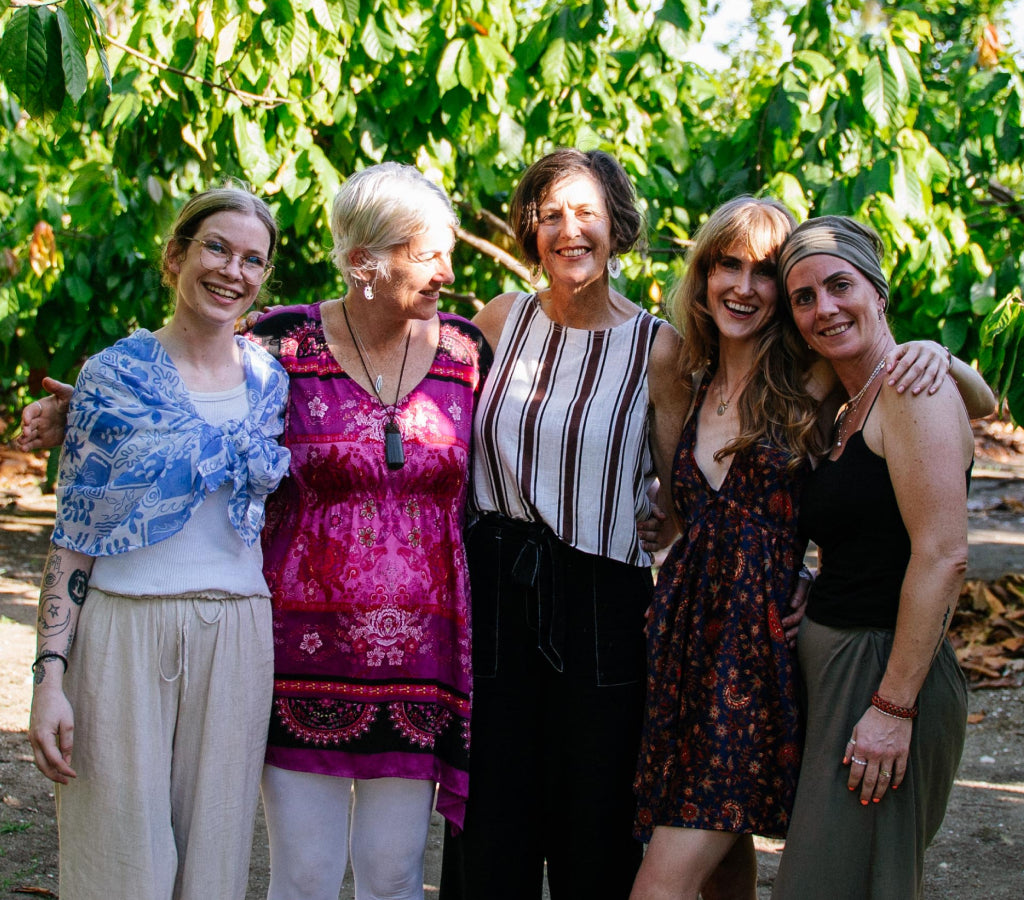Five women posing together outdoors on a cacao farm with greenery in the background