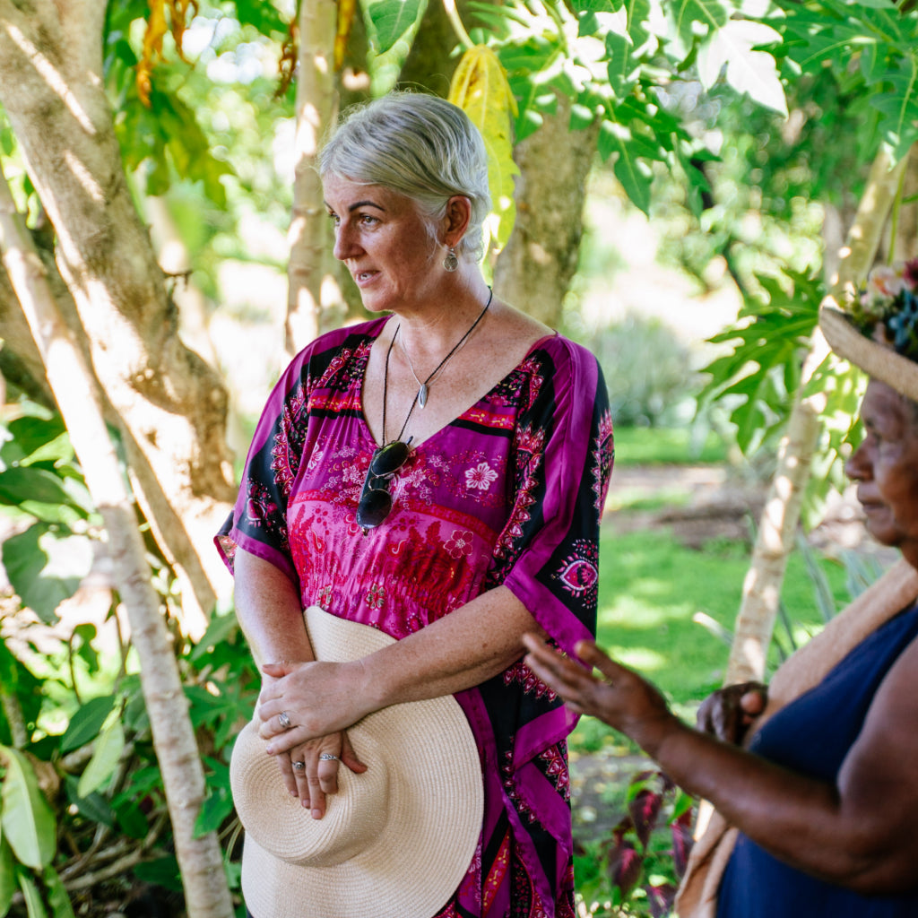 Two women standing in a lush, green outdoor setting with trees and plants at a cacao farm.